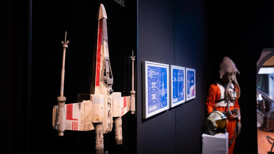 Das Bild zeigt von links nach rechts ein rot-weißes Raumschiff, drei Bilder, die an der Wand hängen und eine Figur im roten Kostüm.