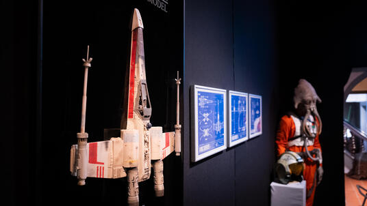 Das Bild zeigt von links nach rechts ein rot-weißes Raumschiff, drei Bilder, die an der Wand hängen und eine Figur im roten Kostüm.