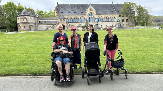 Die Stadtbehindertenbeauftragte Andrea Simon steht mit vier Frauen vor der Wiese der Kaiserpfalz, die im Hintergrund zu sehen ist. Von den vier Frauen sitzt eine Frau im Rollstuhl, eine steht am Rollator und eine schiebt einen Kinderwagen.