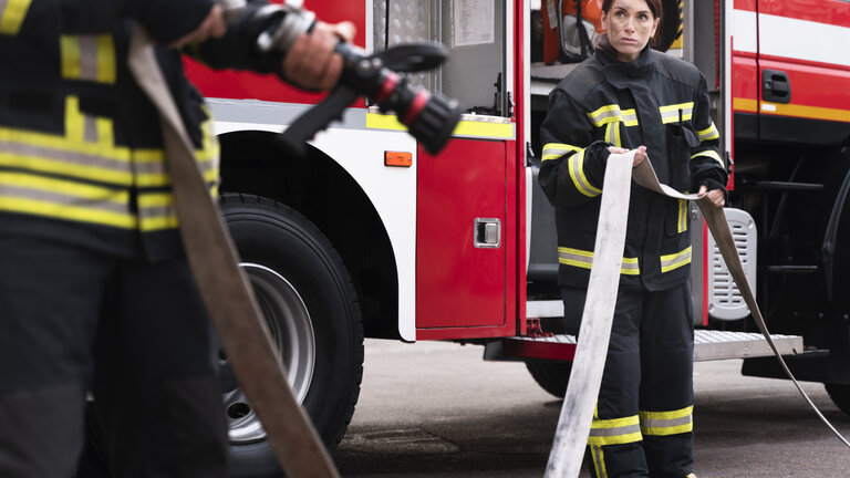 Eine Frau und ein Mann halten einen Feuerwehrschlauch in der Hand neben einem Fahrzeug